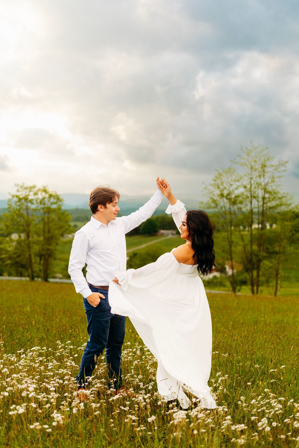 Groom twirling bride in white dress through wildflower meadow with mountain backdrop at golden hour.