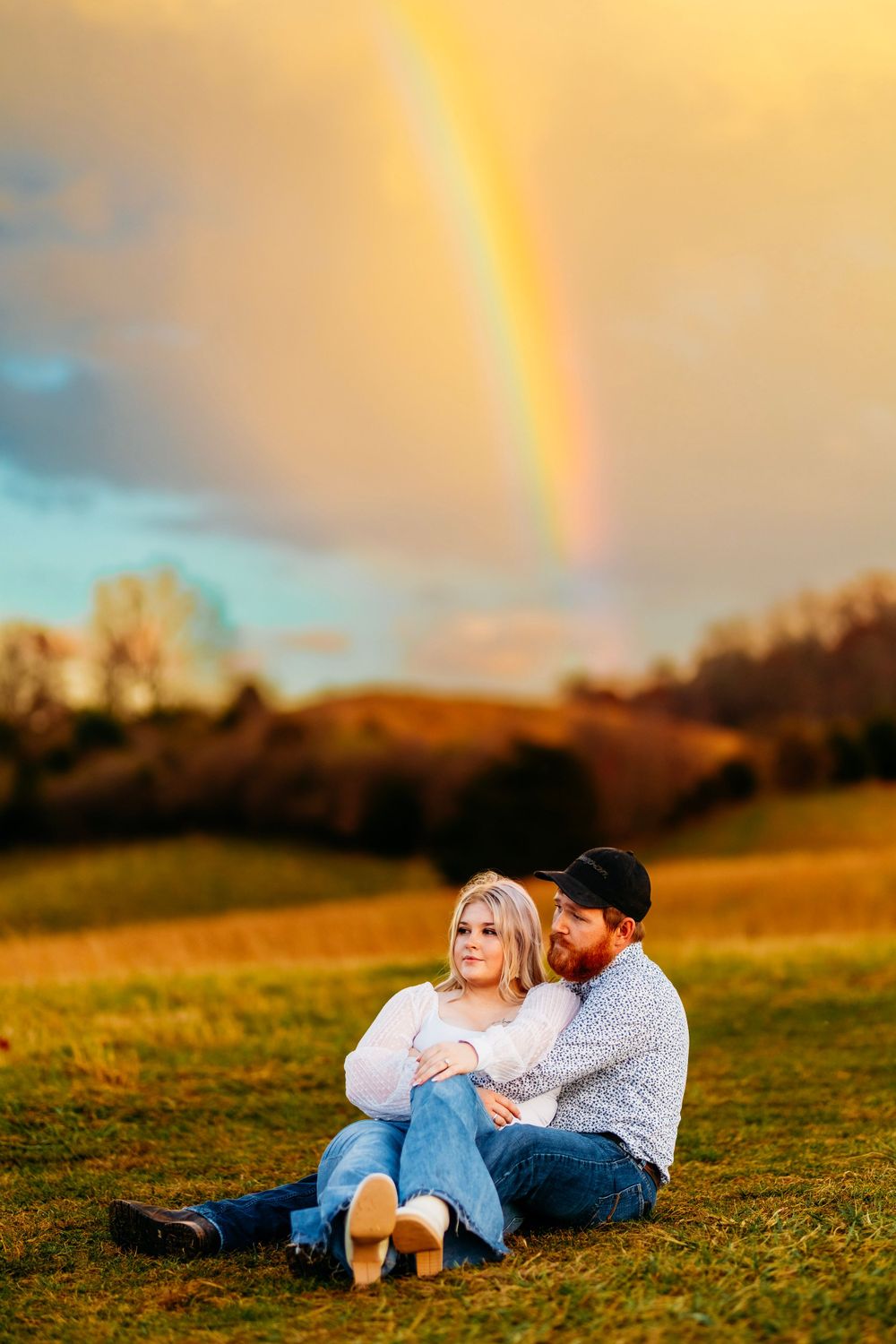 Couple sitting together on a grassy field at sunset with a brilliant rainbow in the sky above.