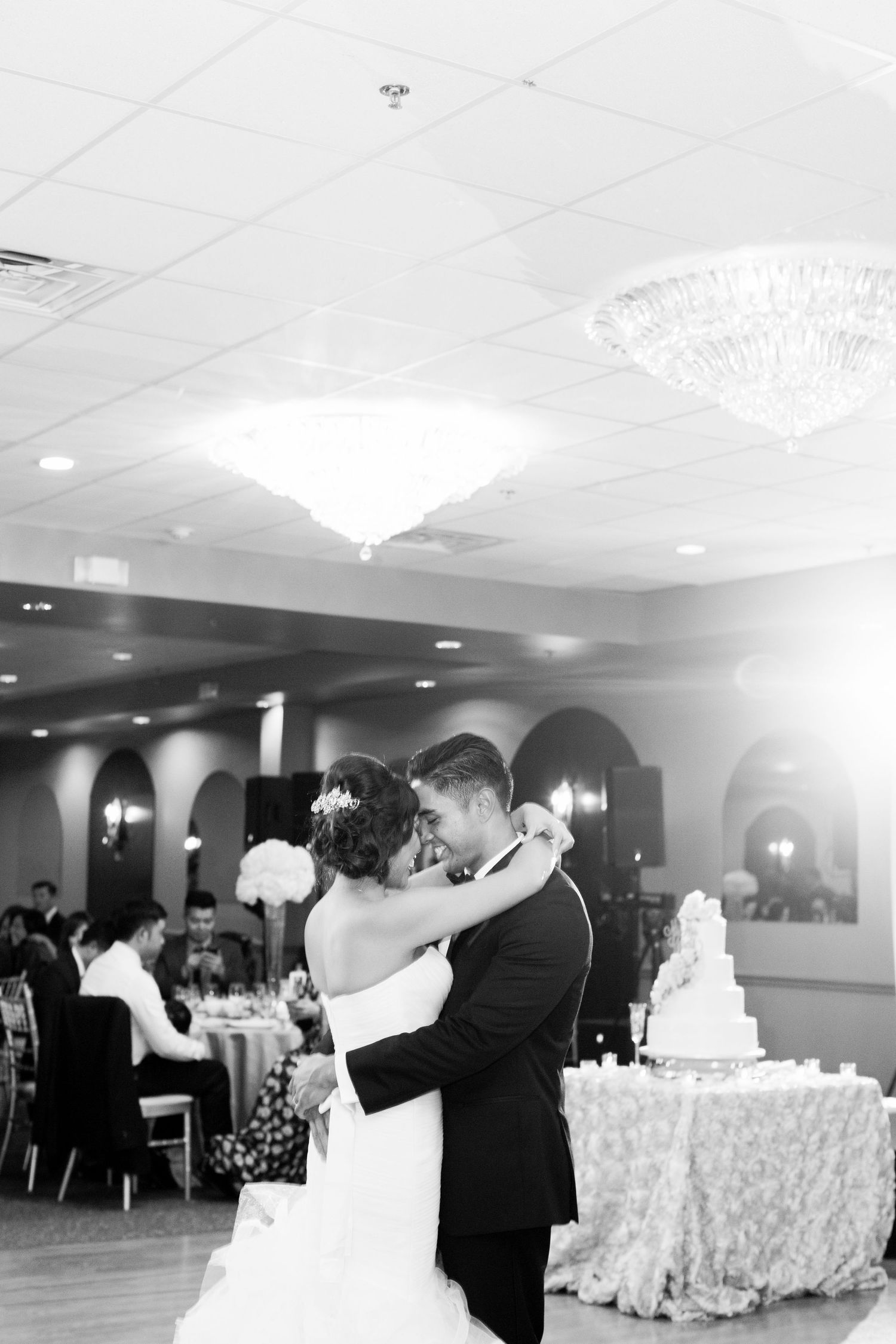 Newlyweds share their first dance in an elegant ballroom with chandelier and wedding cake.
