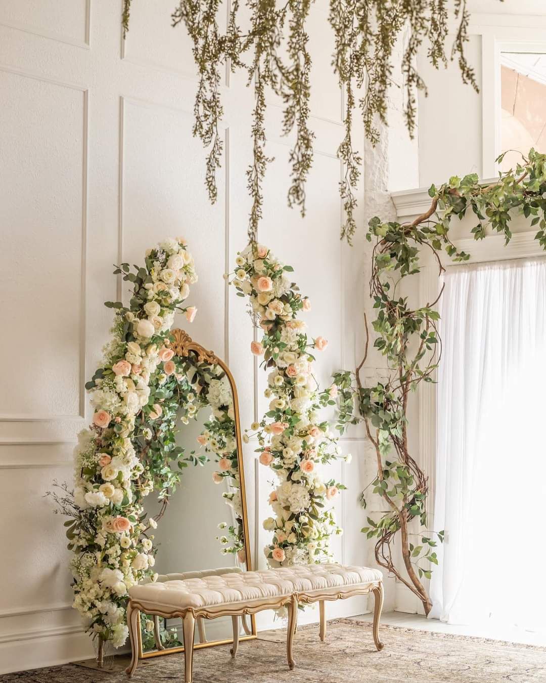 Romantic floral arch installation with hanging greenery and cream roses against white paneled wall.