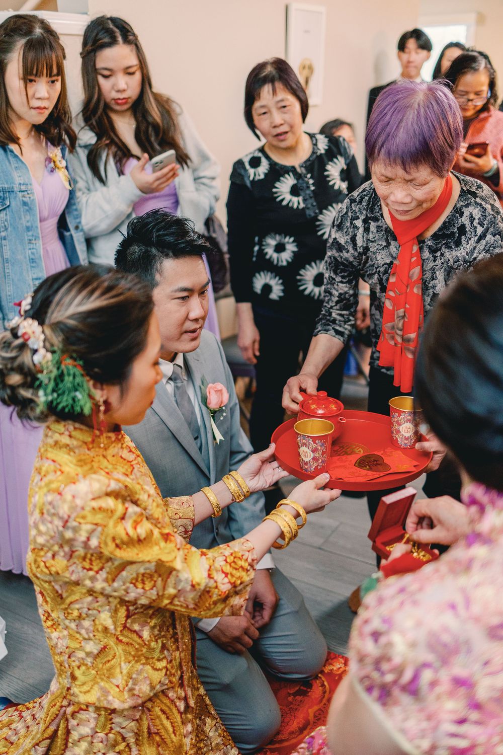 Chinese wedding tea ceremony with bride in gold qipao and groom in gray suit kneeling to serve tea to elders.