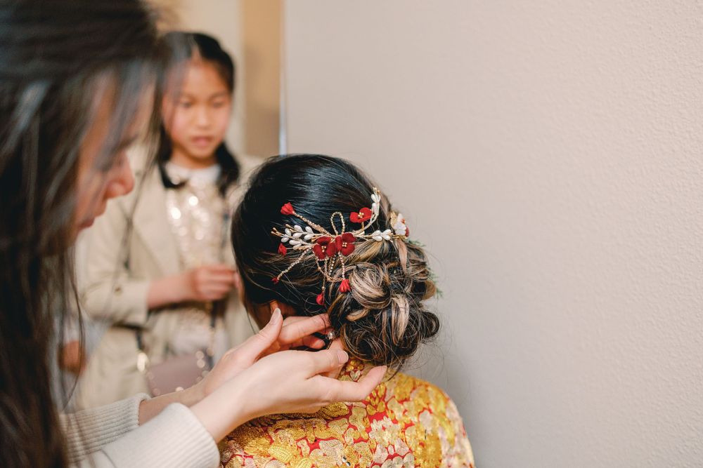 Hairstylist arranging elegant red floral hair comb in bride's dark updo, with young girl watching in background.