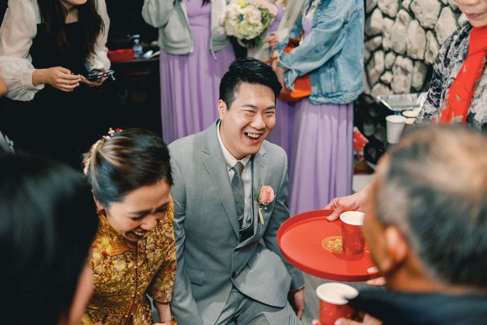 Groom laughing joyfully during a traditional Chinese tea ceremony, holding a red tray with guests in festive attire.