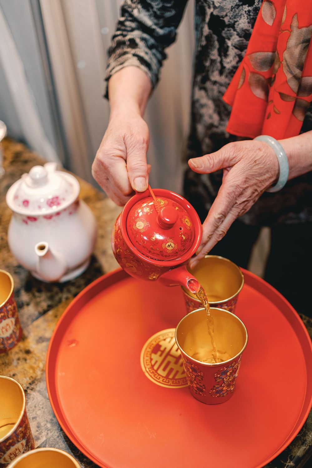 Elderly woman pouring tea from a red Chinese teapot into gold cups on a red tray during a traditional tea ceremony.