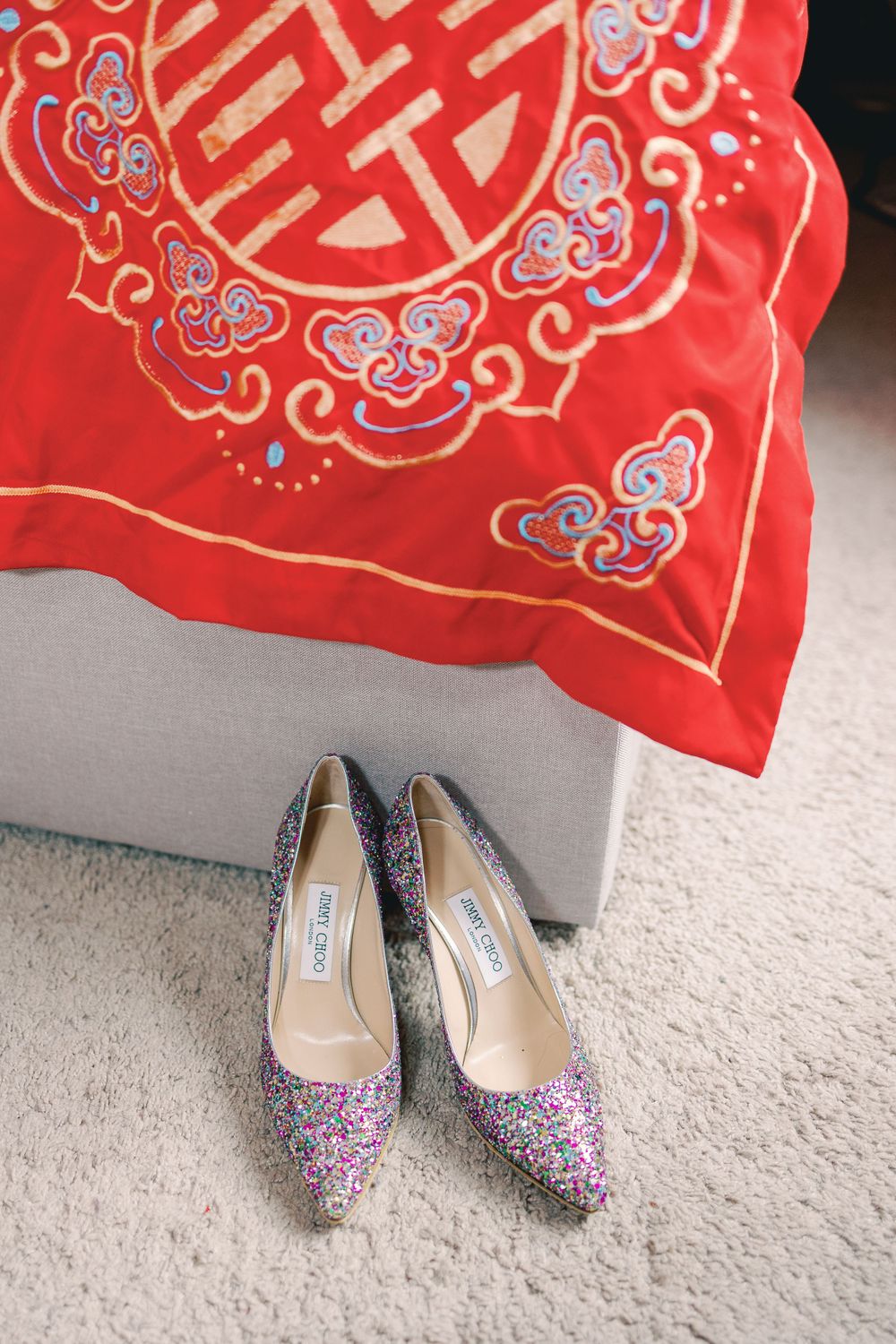 Glittery Jimmy Choo heels on carpet beside a red embroidered Chinese double happiness cushion for a wedding.