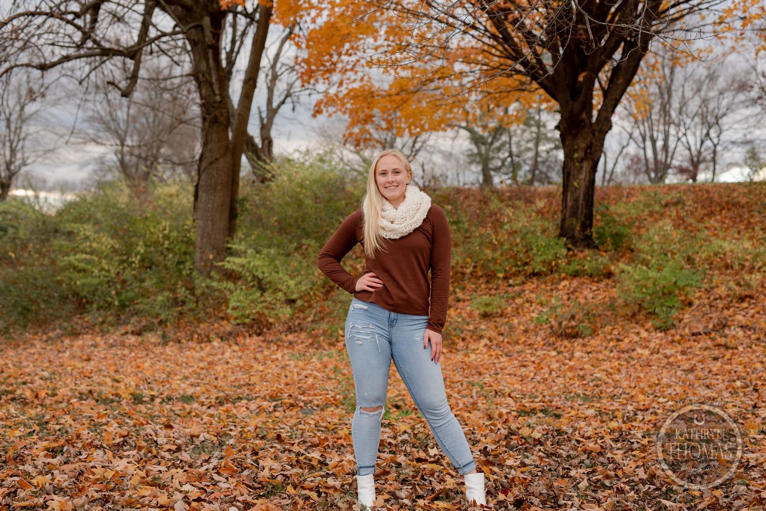 Person in brown sweater and jeans standing among fall foliage with orange trees in background.