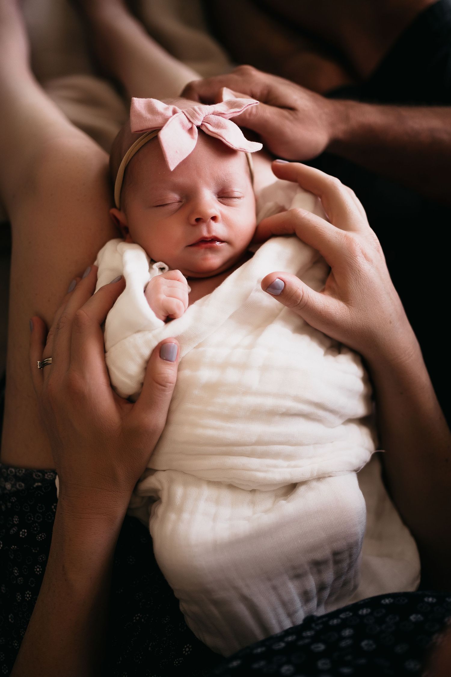 A sleeping newborn baby wearing a pink bow headband wrapped in white fabric.