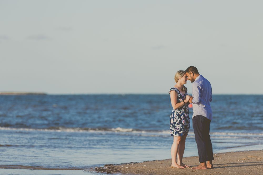 Couple standing close together on beach with waves and horizon in background.