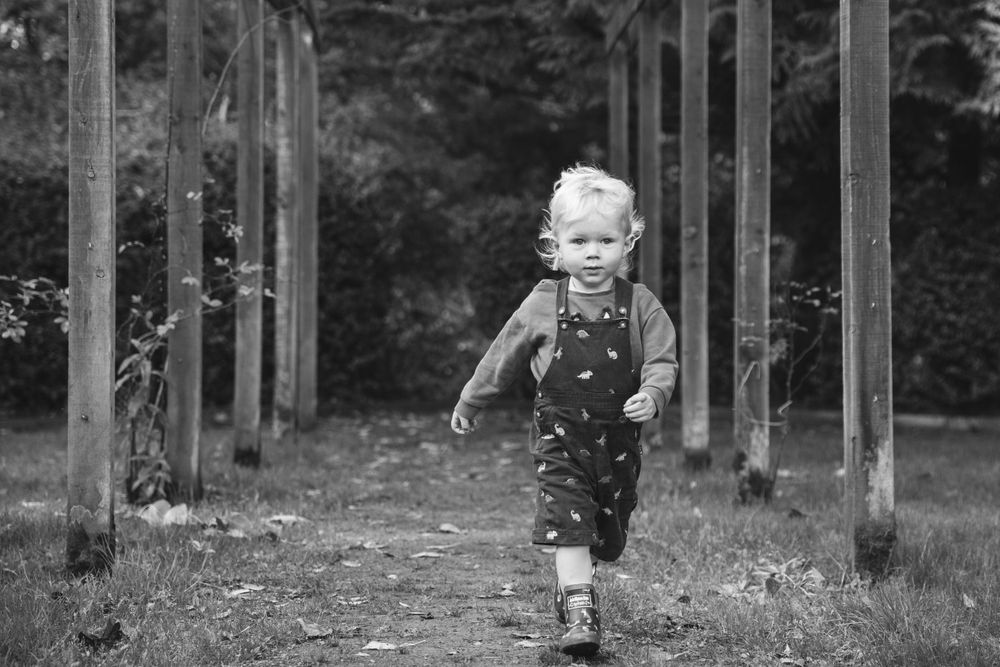 Black and white photo of toddler in overalls running through garden posts.
