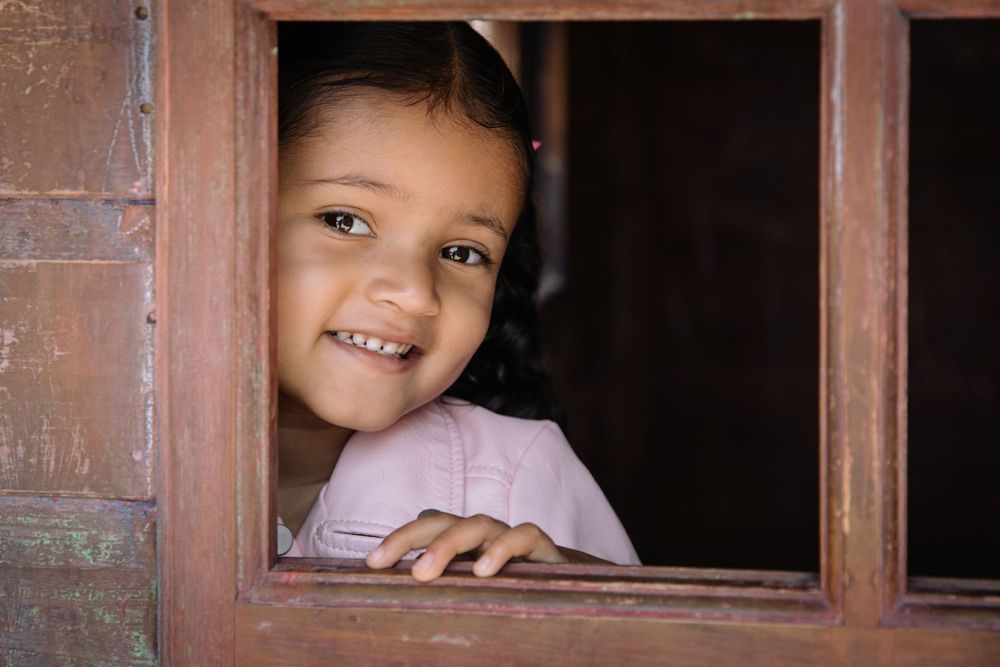 Smiling child peeks playfully through a wooden window frame in natural lighting.