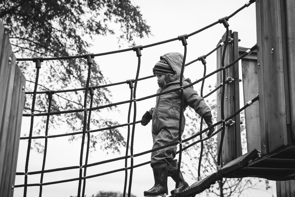 Small child in winter clothing carefully crosses a rope bridge at an outdoor playground in black and white.