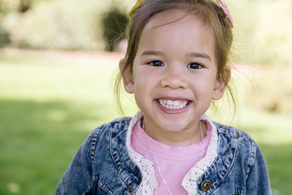 Child wearing pink shirt and denim jacket smiles brightly outdoors.