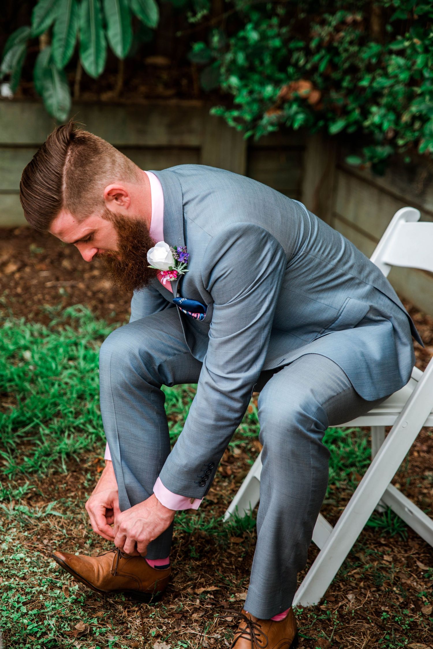 A groom in a grey suit ties his brown leather shoes while seated on a white folding chair.