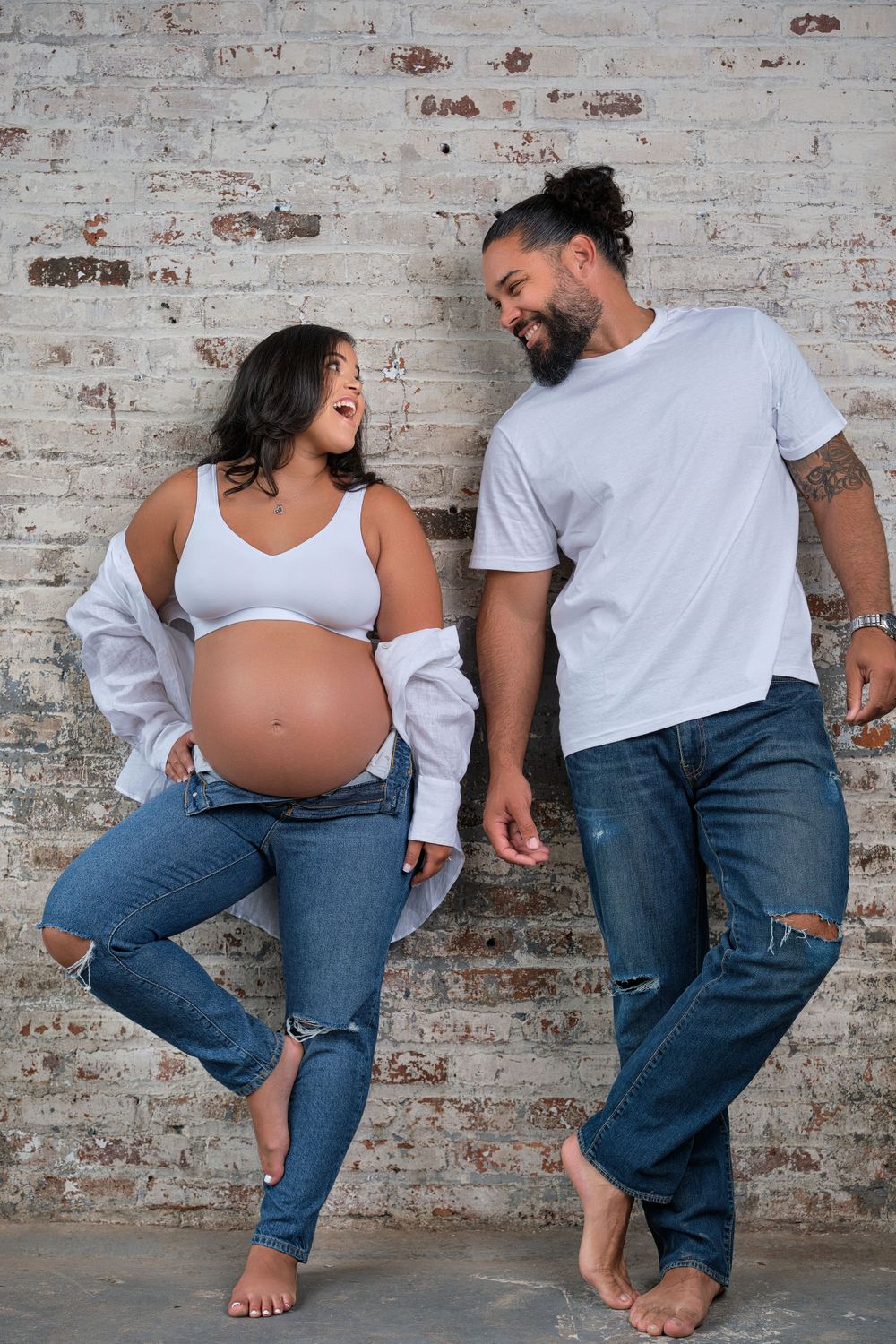 Candid maternity photo of couple laughing against rustic brick wall background.