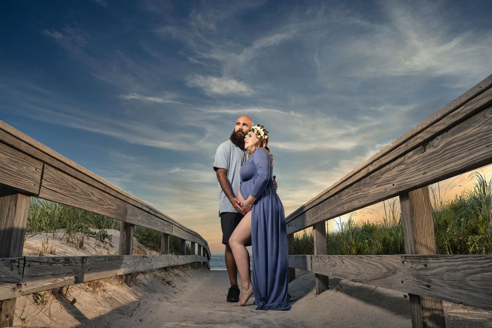 Elegant seaside maternity pose on wooden walkway during sunset.