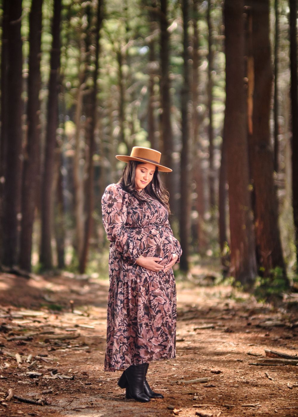 Maternity portrait of woman in floral dress and hat standing on wooded trail.