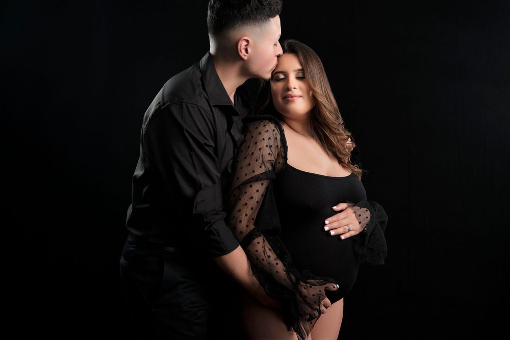 Intimate maternity photography session against dark background with couple in black attire.