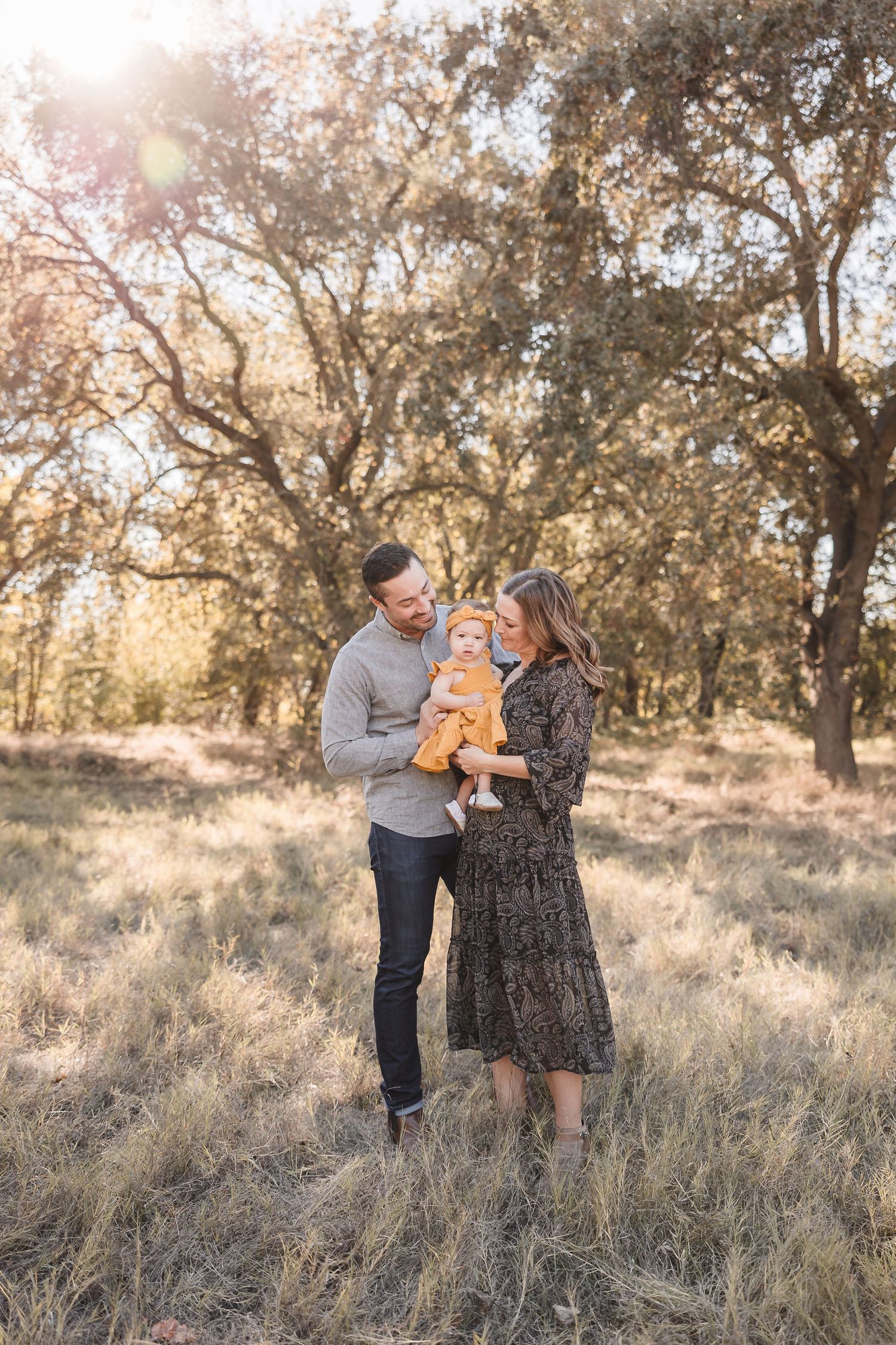 A couple holds their young child in a sunlit field with oak trees in the background.