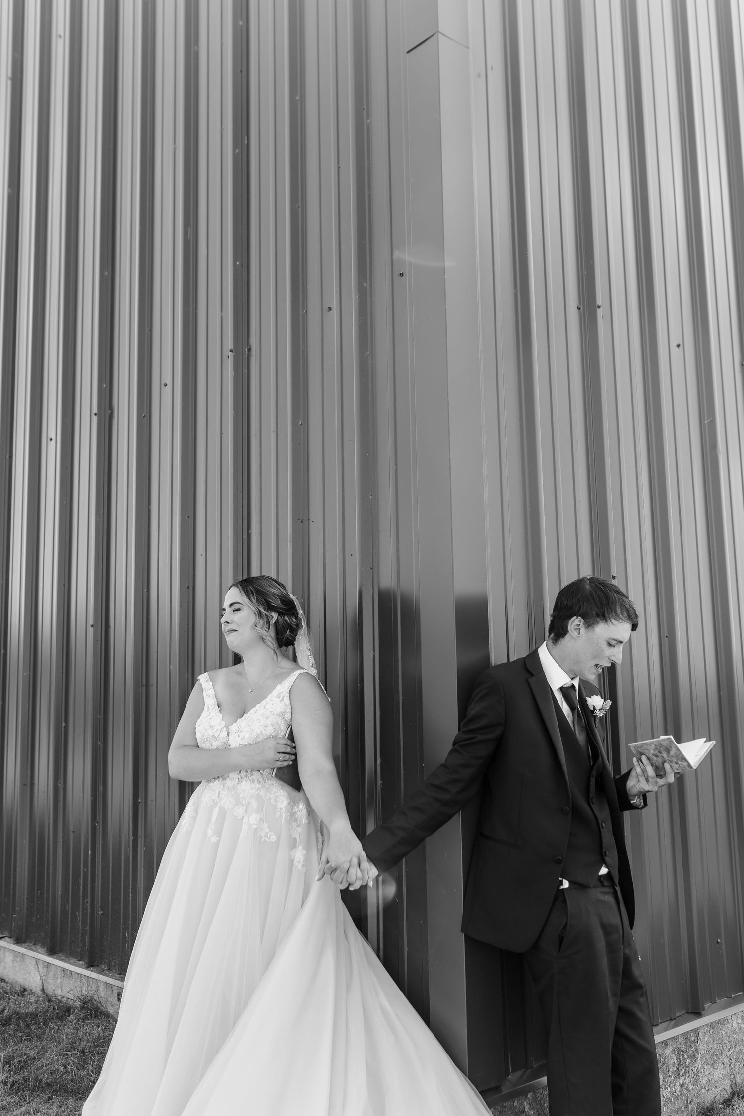 Black and white photo of couple holding hands during first look against industrial wall.