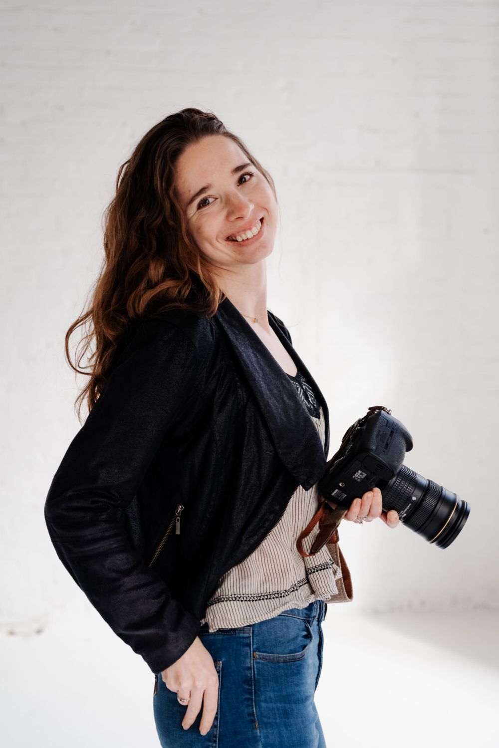 The photographer poses with her camera in a studio with white walls.