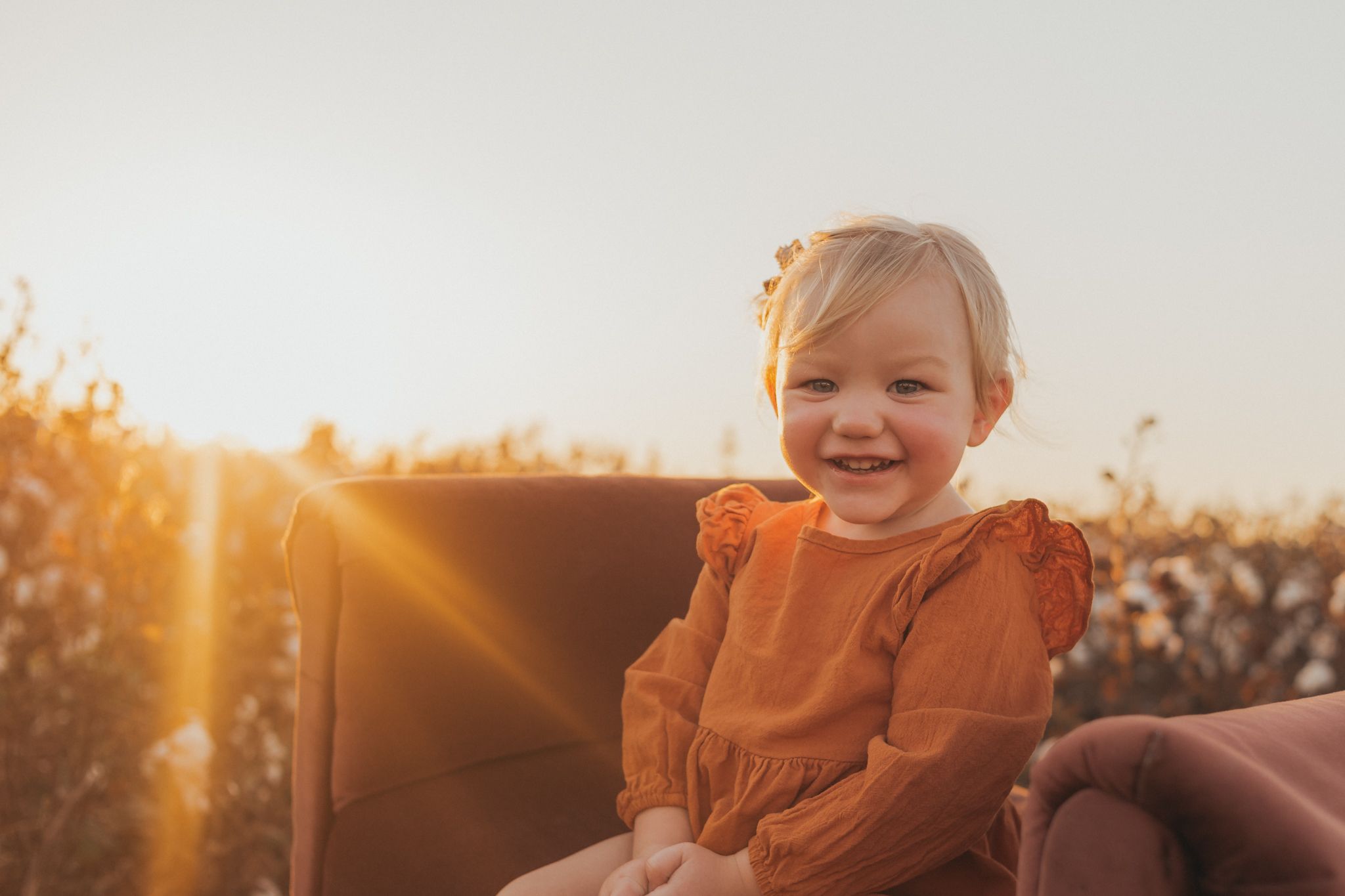 Child in orange dress sitting in brown chair during sunset with sun flare.