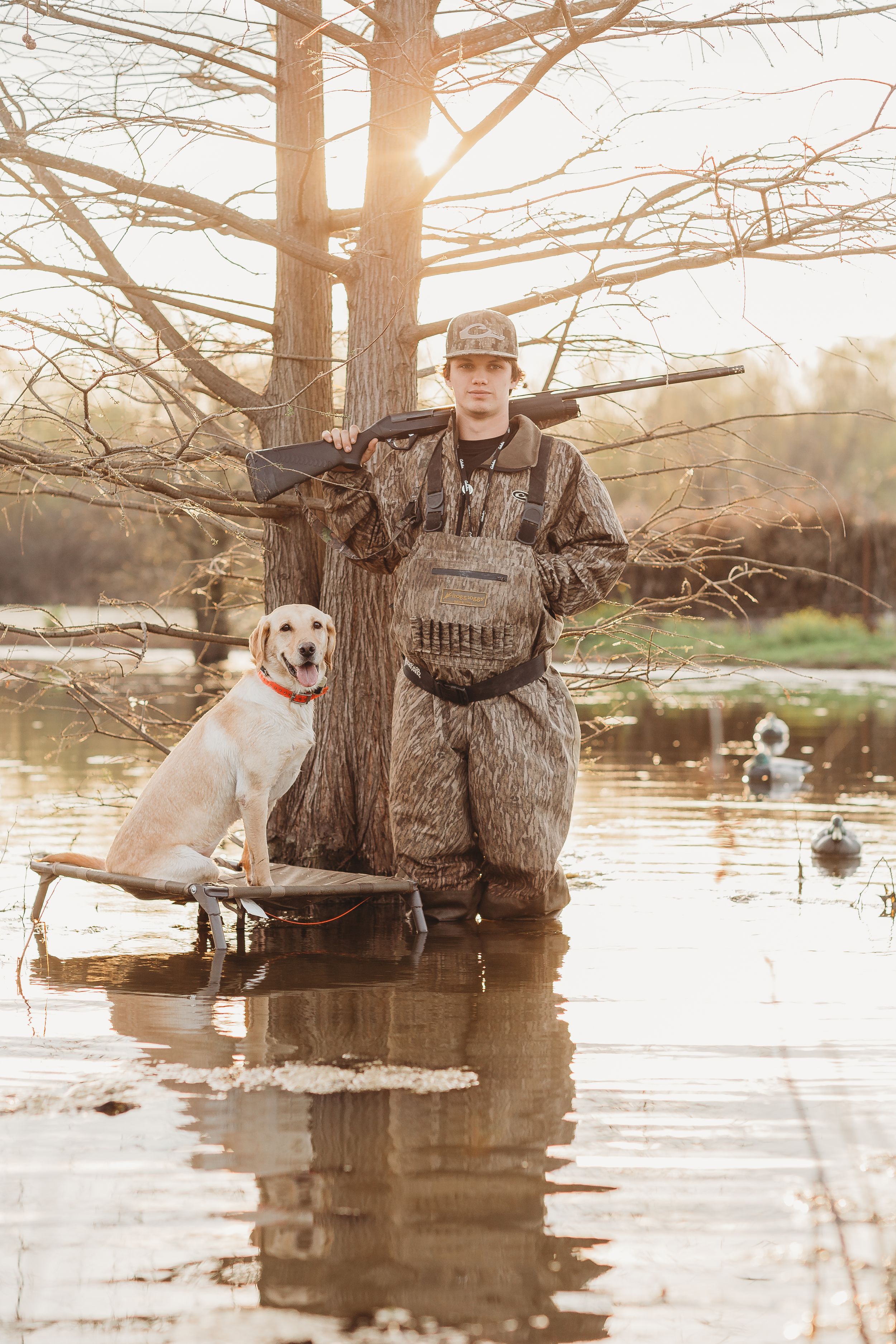 Duck hunter in camouflage gear standing in water with yellow lab during golden hour.