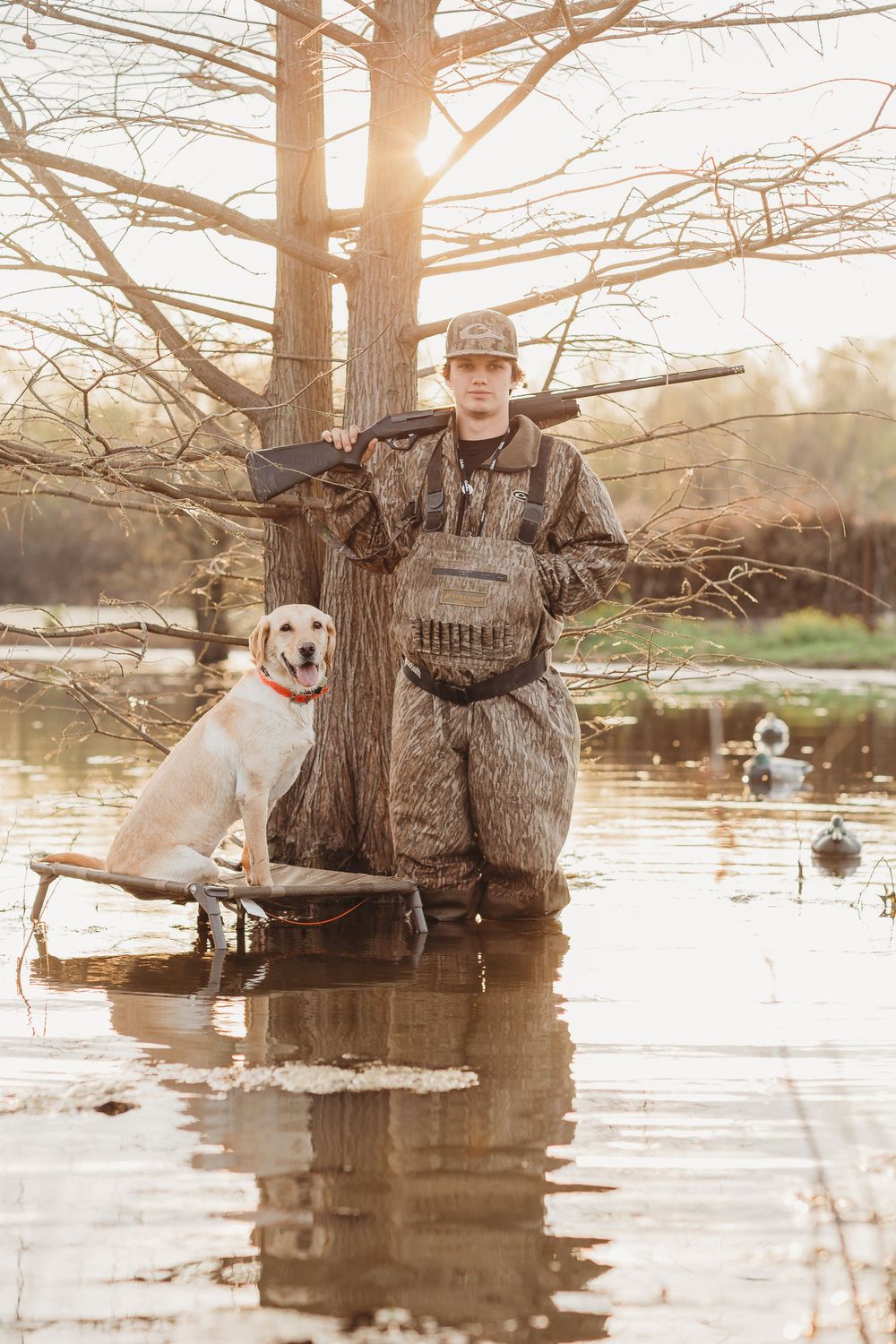 Duck hunter in camouflage gear standing in water with yellow lab during golden hour.