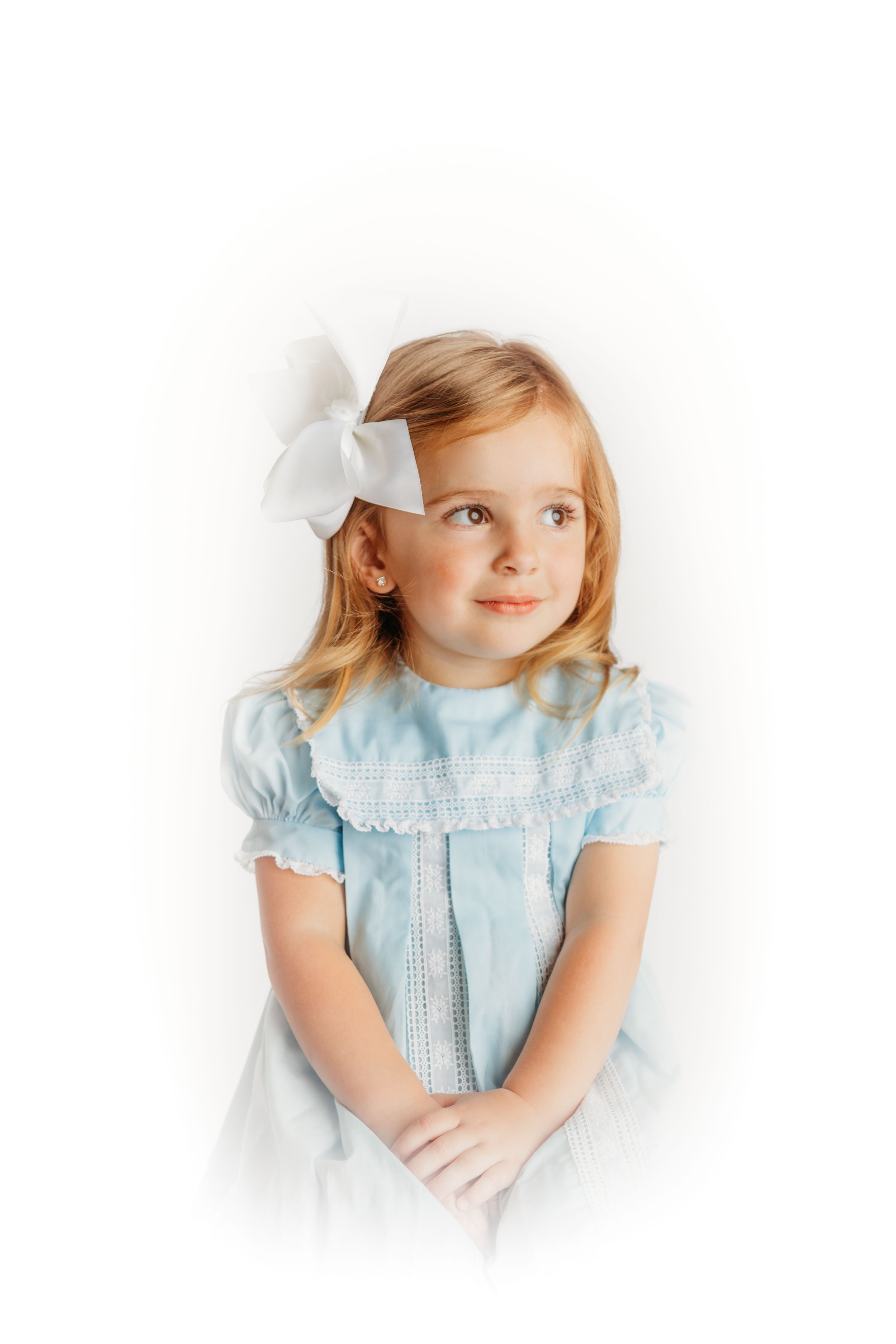 Young child in a light blue vintage-style dress with white bow posed against white background.
