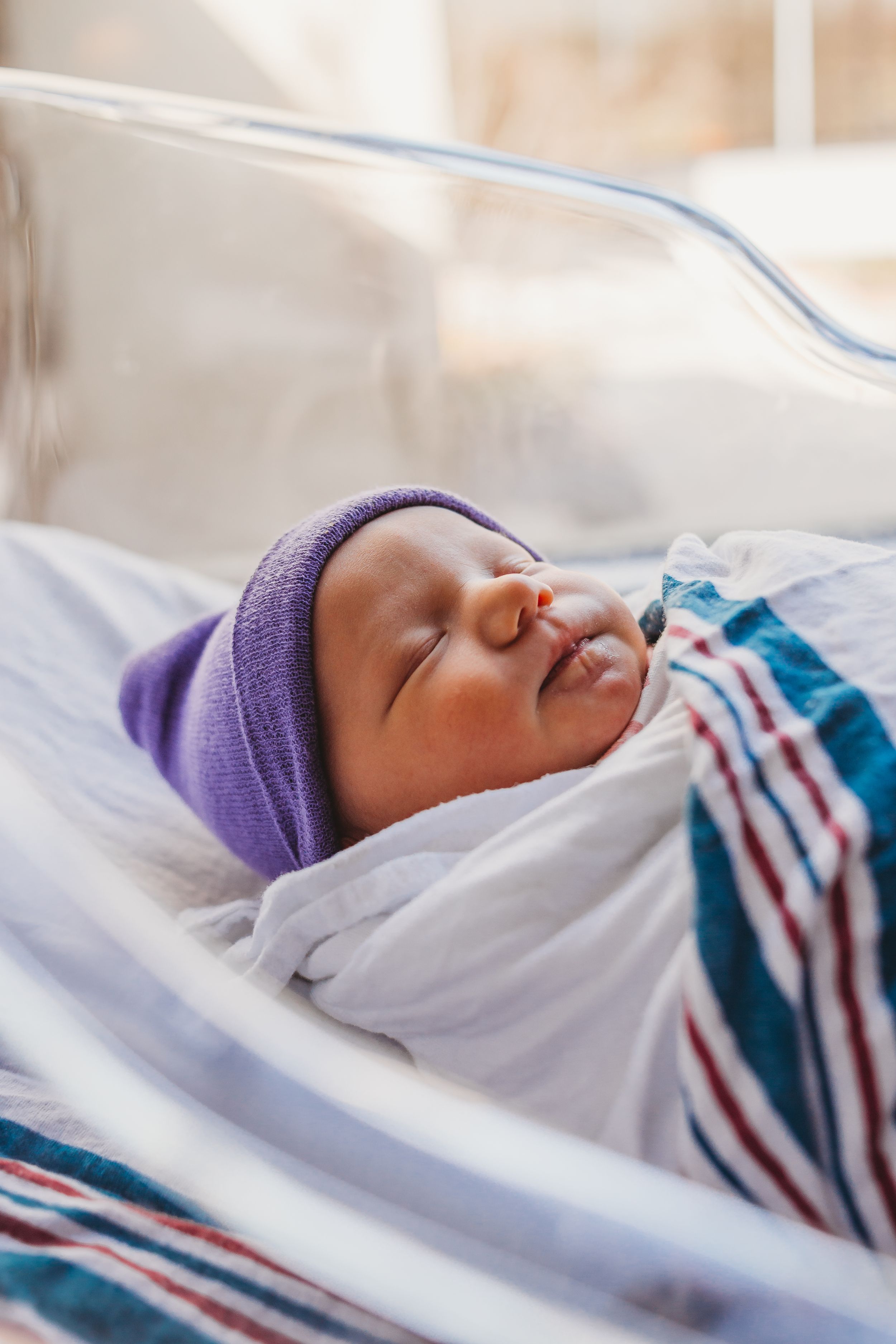 Sleeping newborn wearing purple hat and striped swaddle in hospital bassinet.