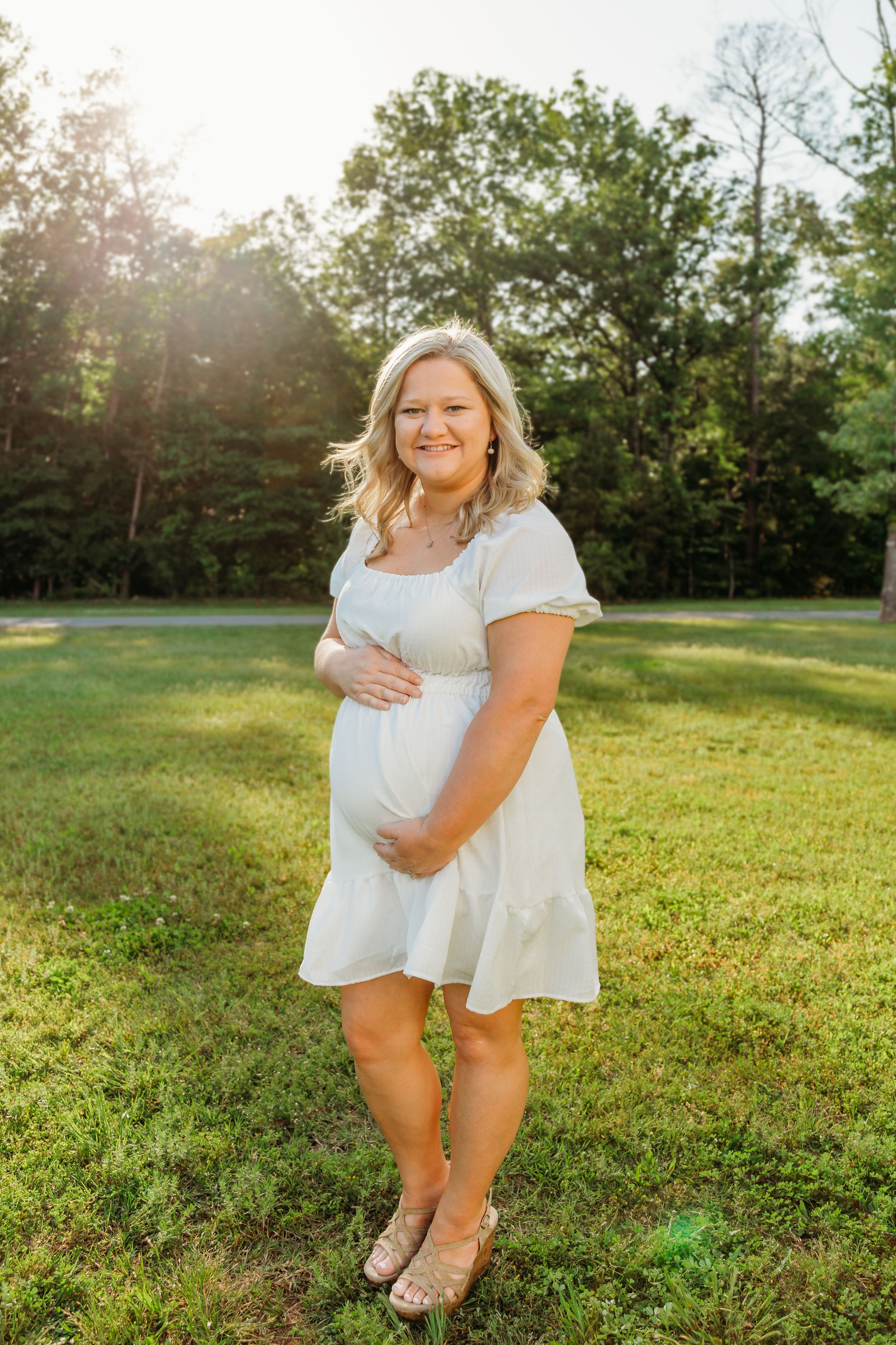 Expectant woman in white dress poses outdoors during sunset in a grassy field.