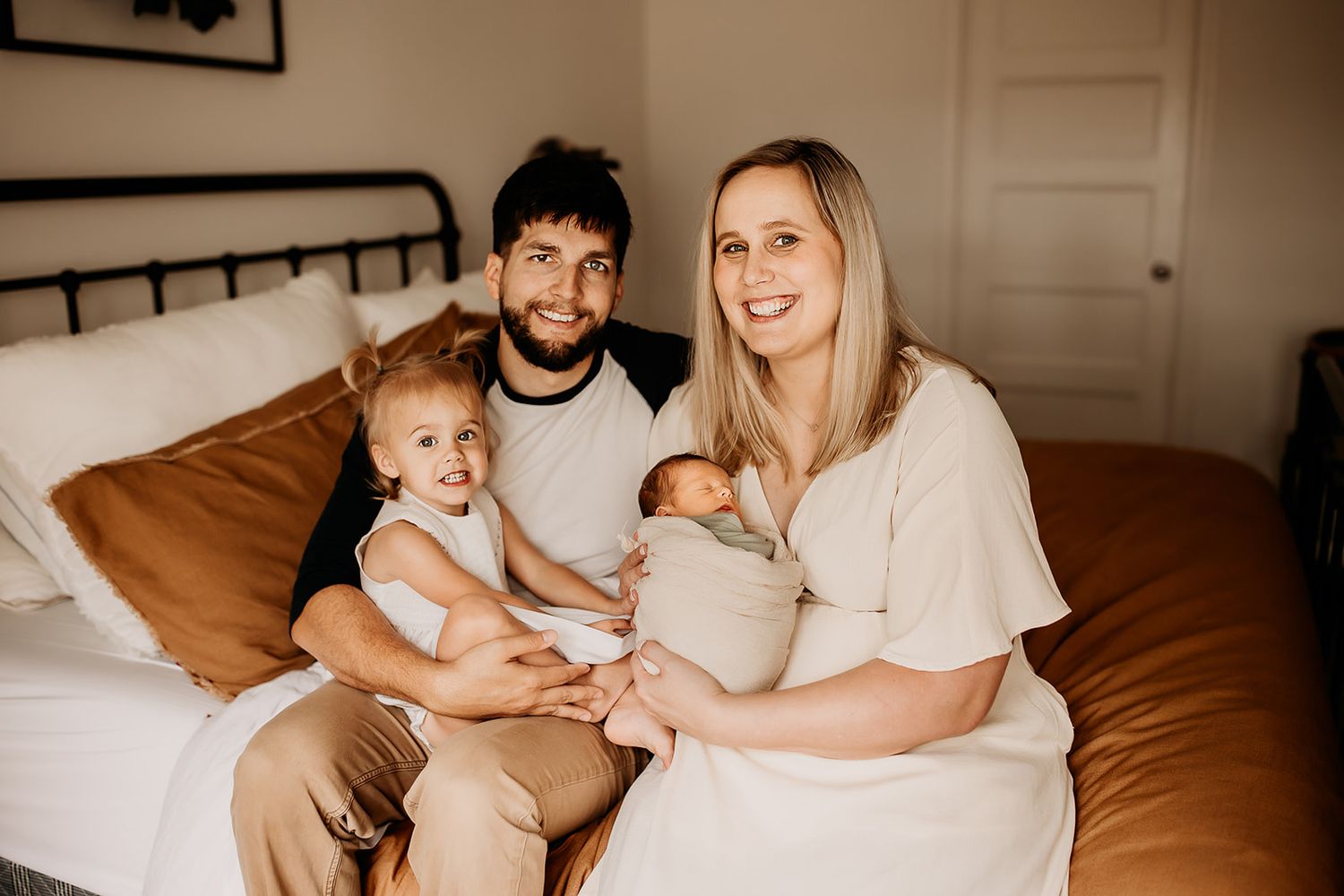 A family of four sits on the bed holding their new baby boy during their newborn photoshoot.