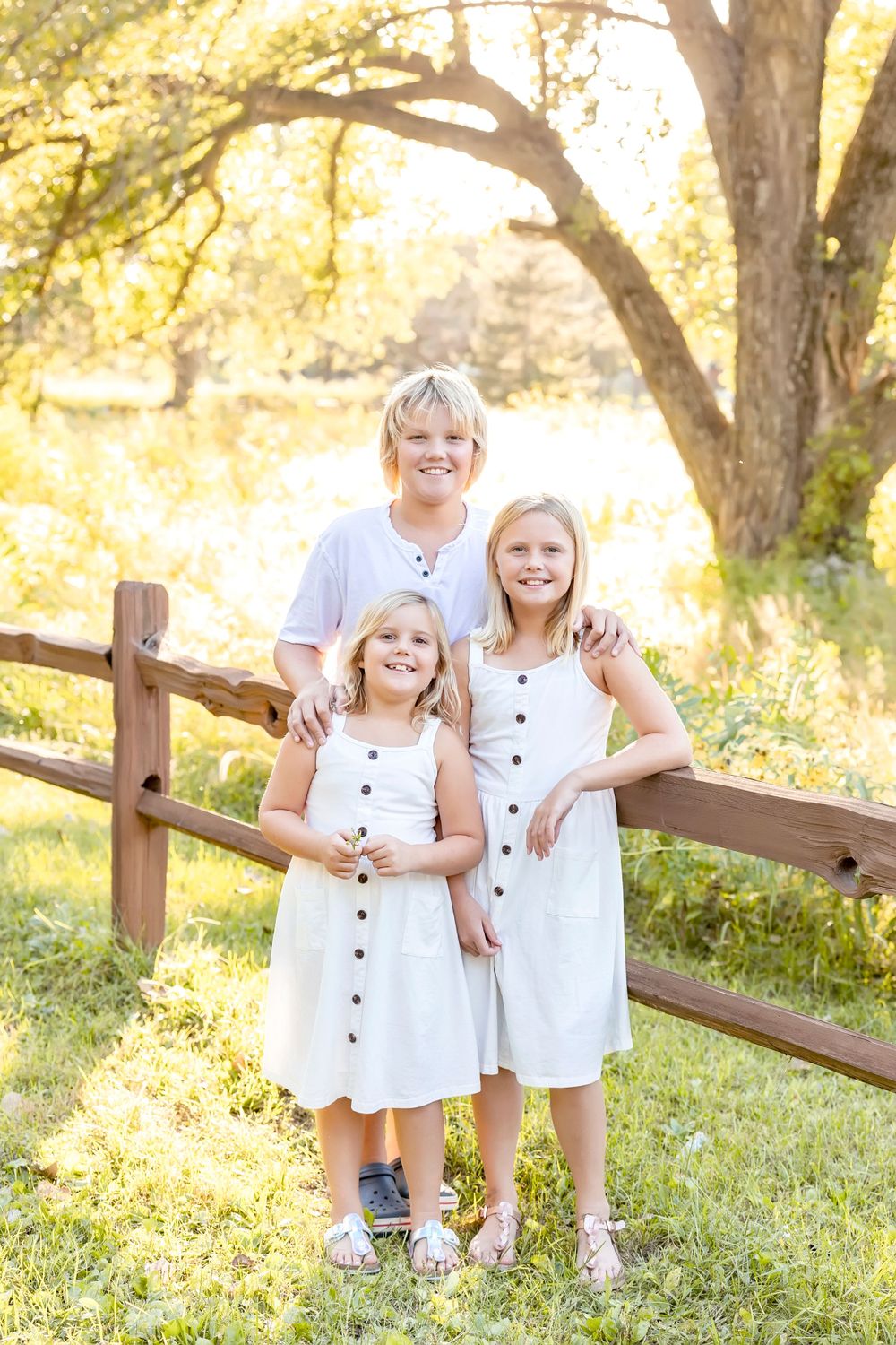 Children in matching white outfits pose along wooden fence during sunset photo session.