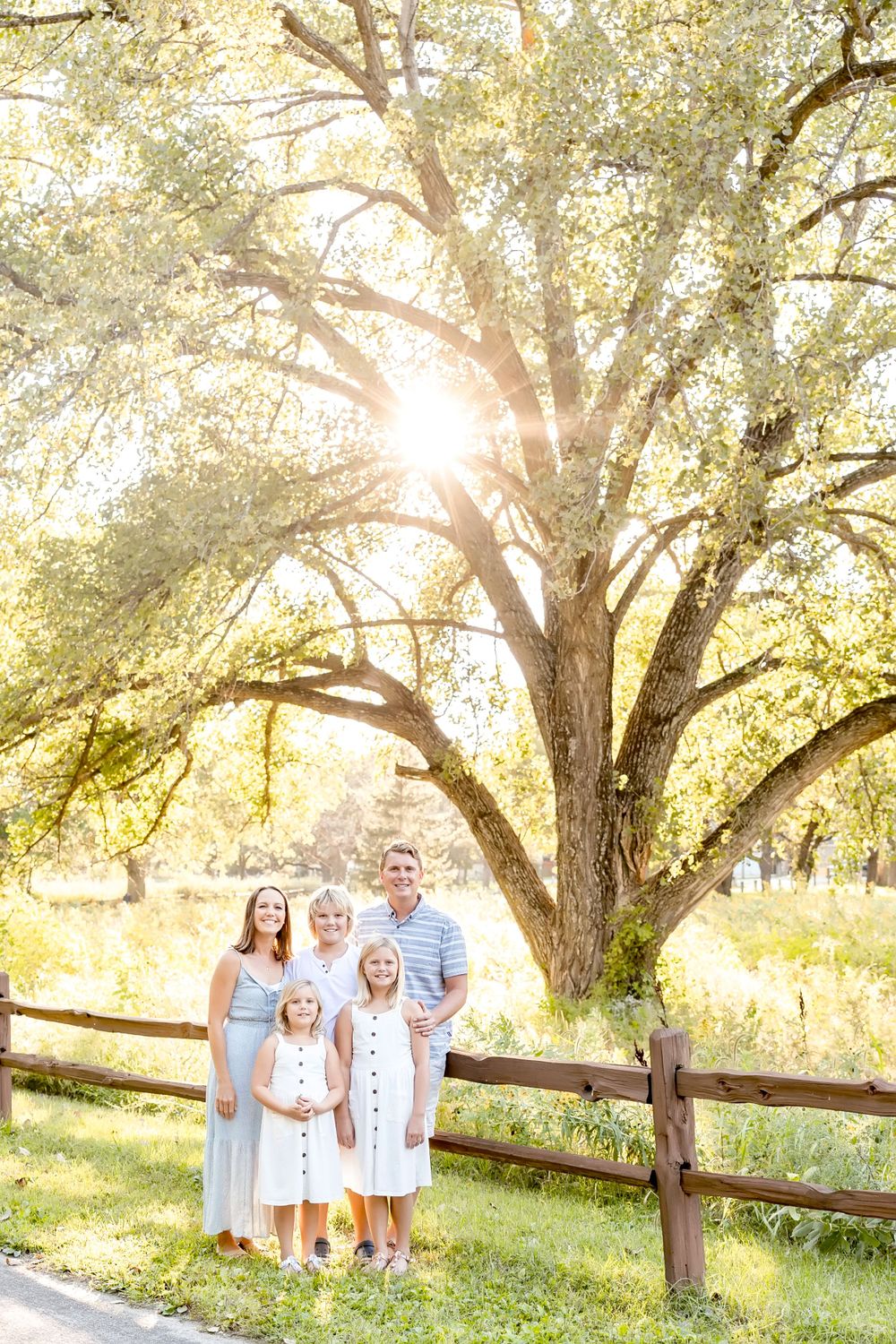A family dressed in white poses together against a backdrop of sunlit trees and a rustic wooden fence.