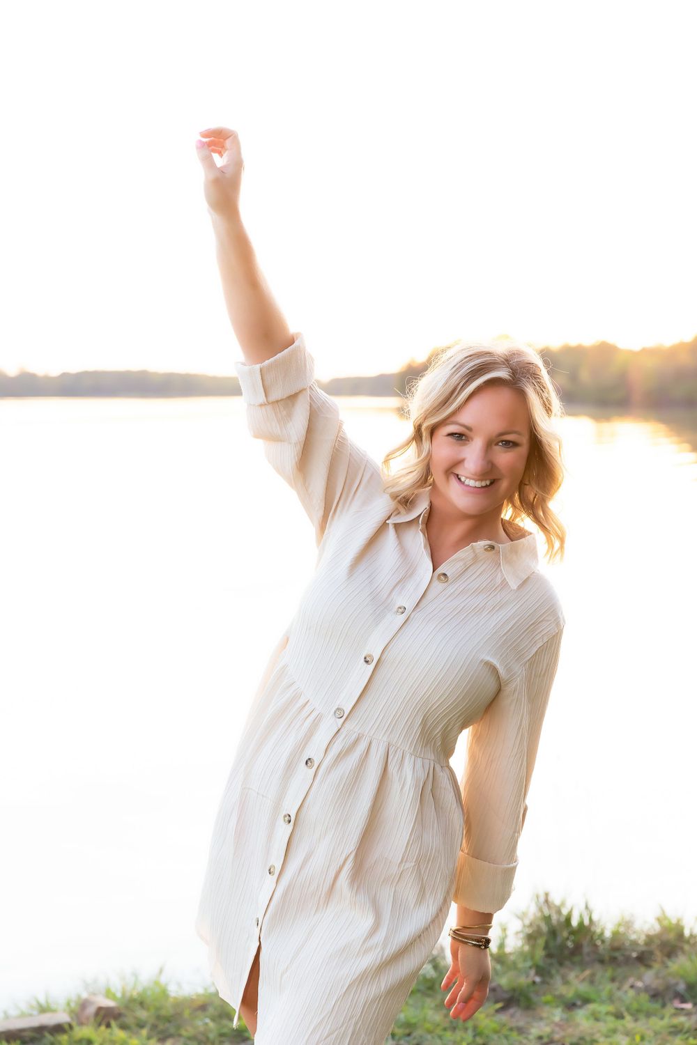 Woman in white button-down dress poses joyfully at sunset by a lake.