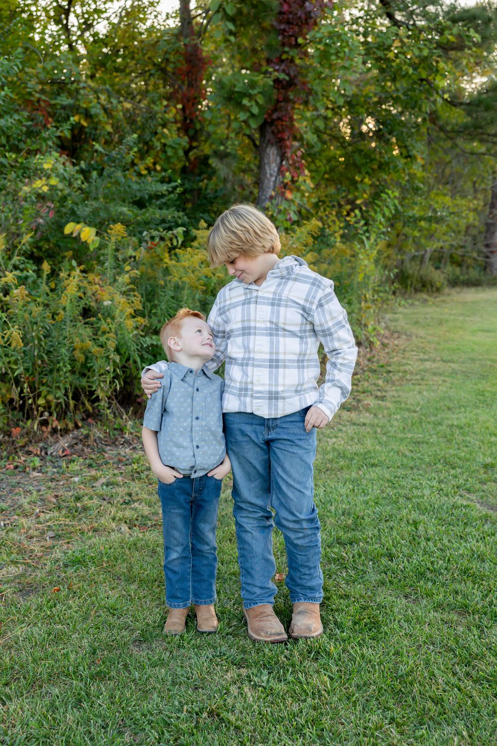 Two young people share an affectionate moment while standing on grass near trees.