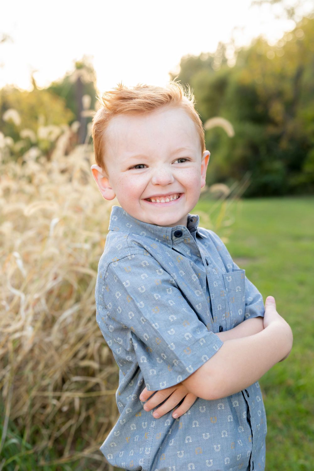 Young child in blue button-up shirt smiling outdoors near ornamental grasses.