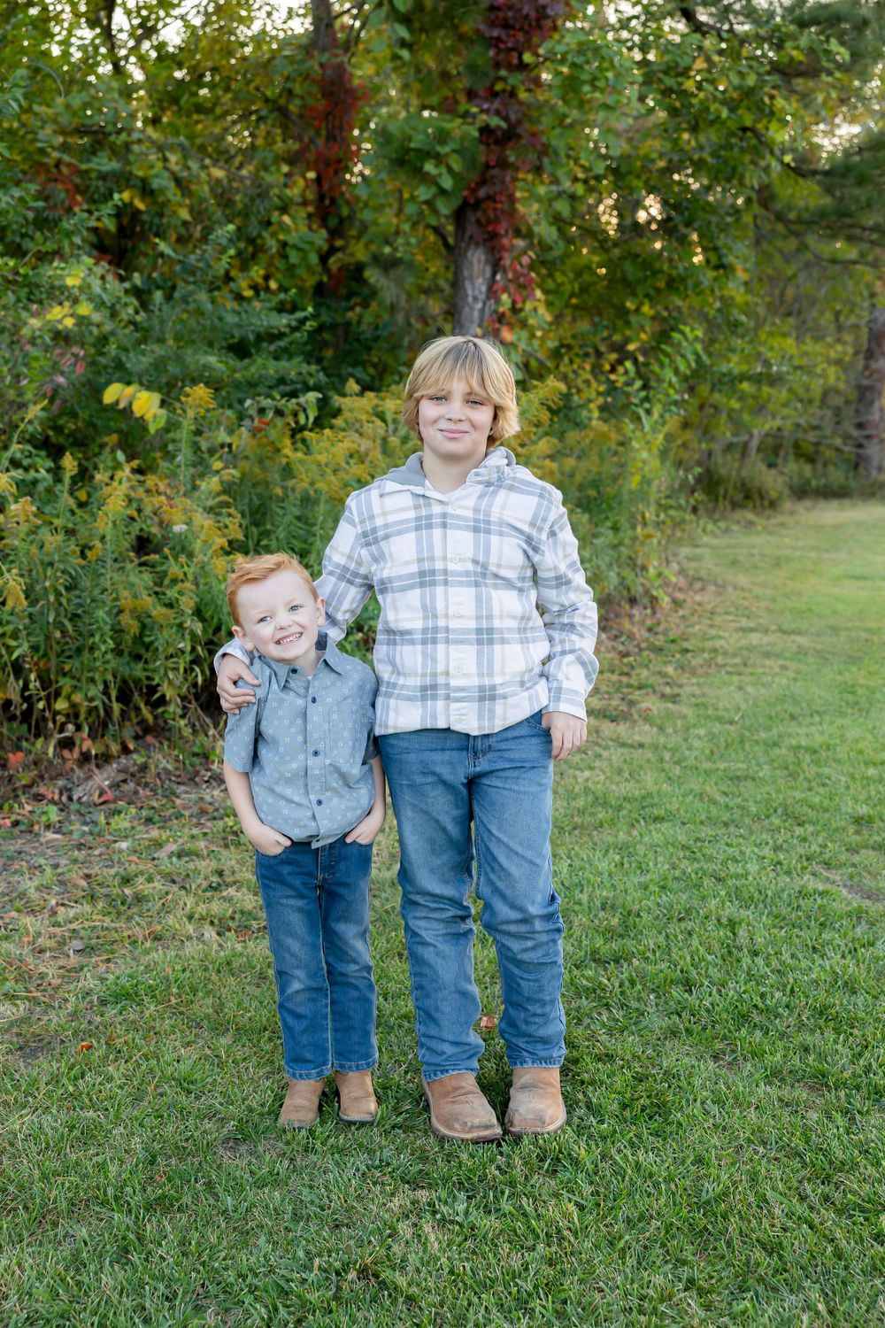 Two young people in plaid shirts stand together on green grass near autumn foliage.