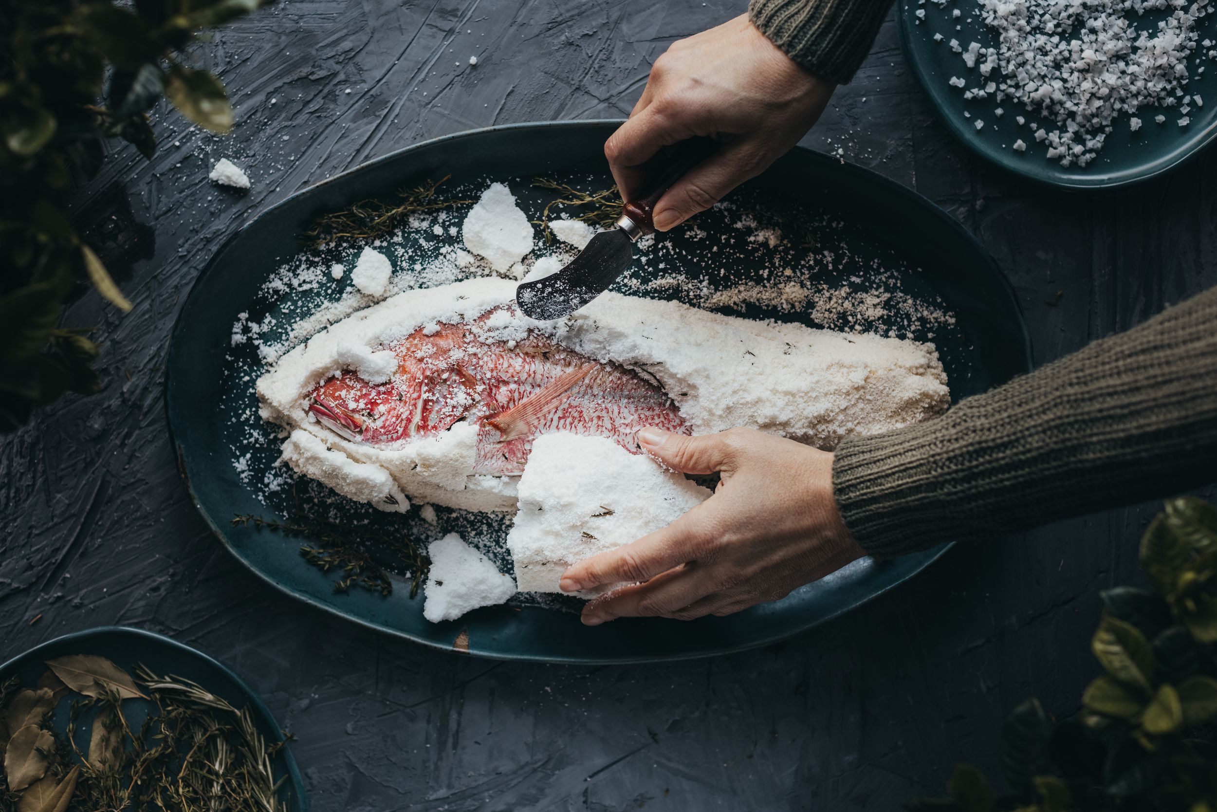 Hands breaking open a salt crust on a baked red fish served on a dark plate.
