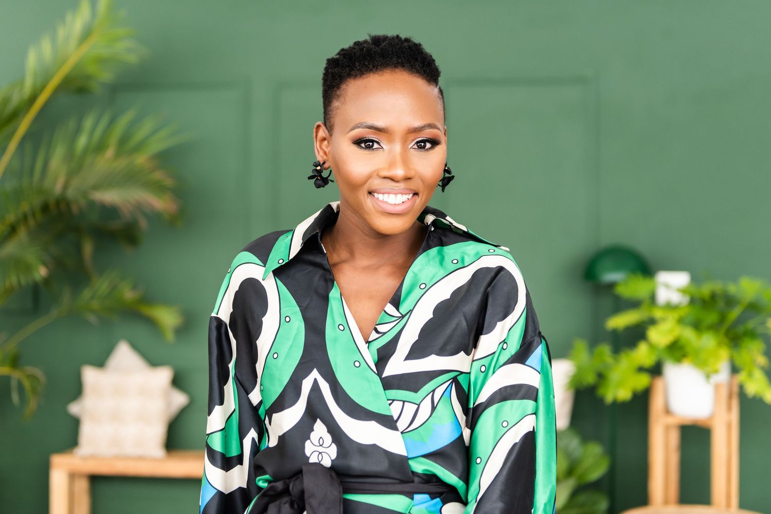 Professional in green patterned top poses against emerald wall with tropical plants.