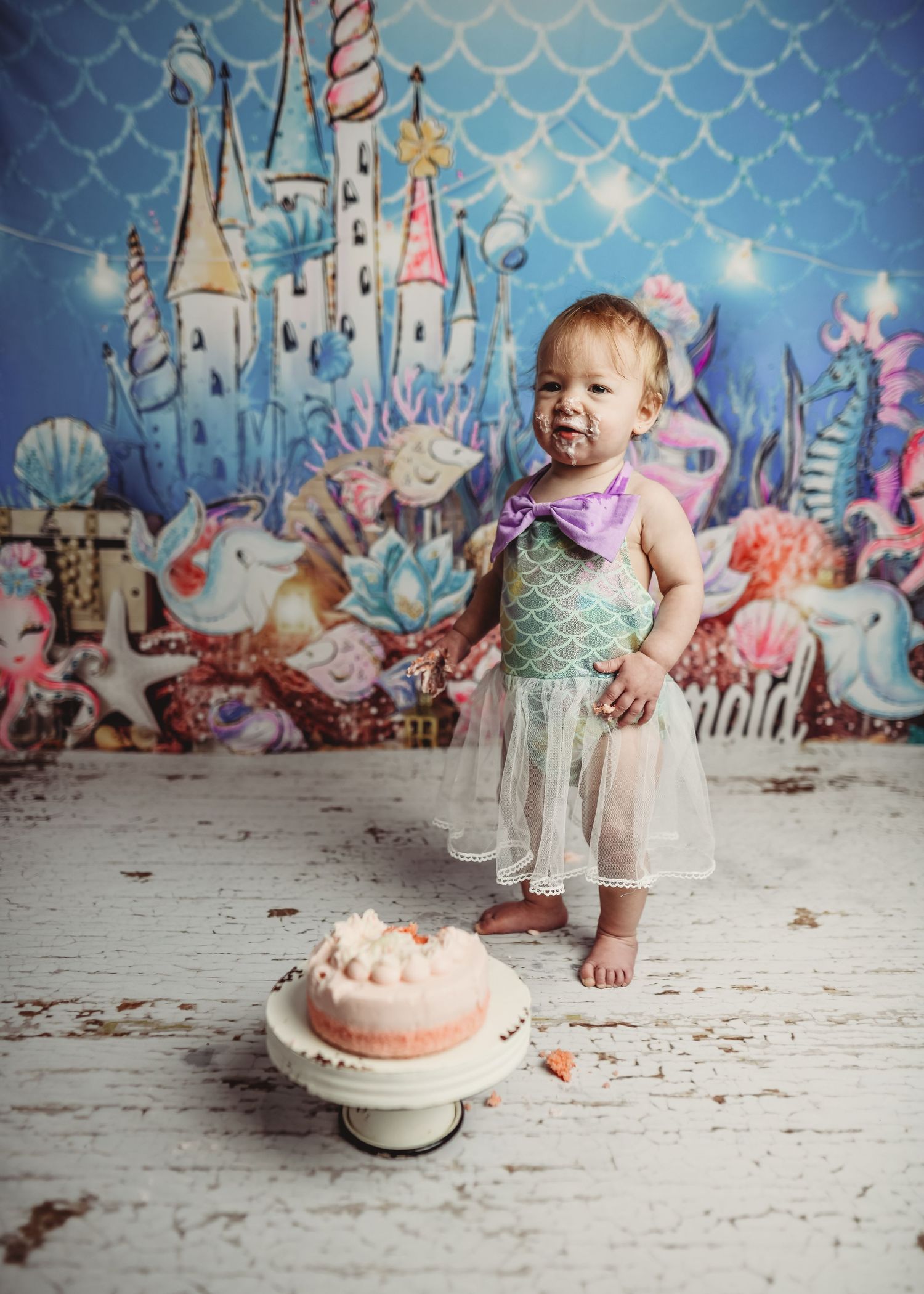 Whimsical mermaid-themed backdrop with cake and pastel decorations on white floor.