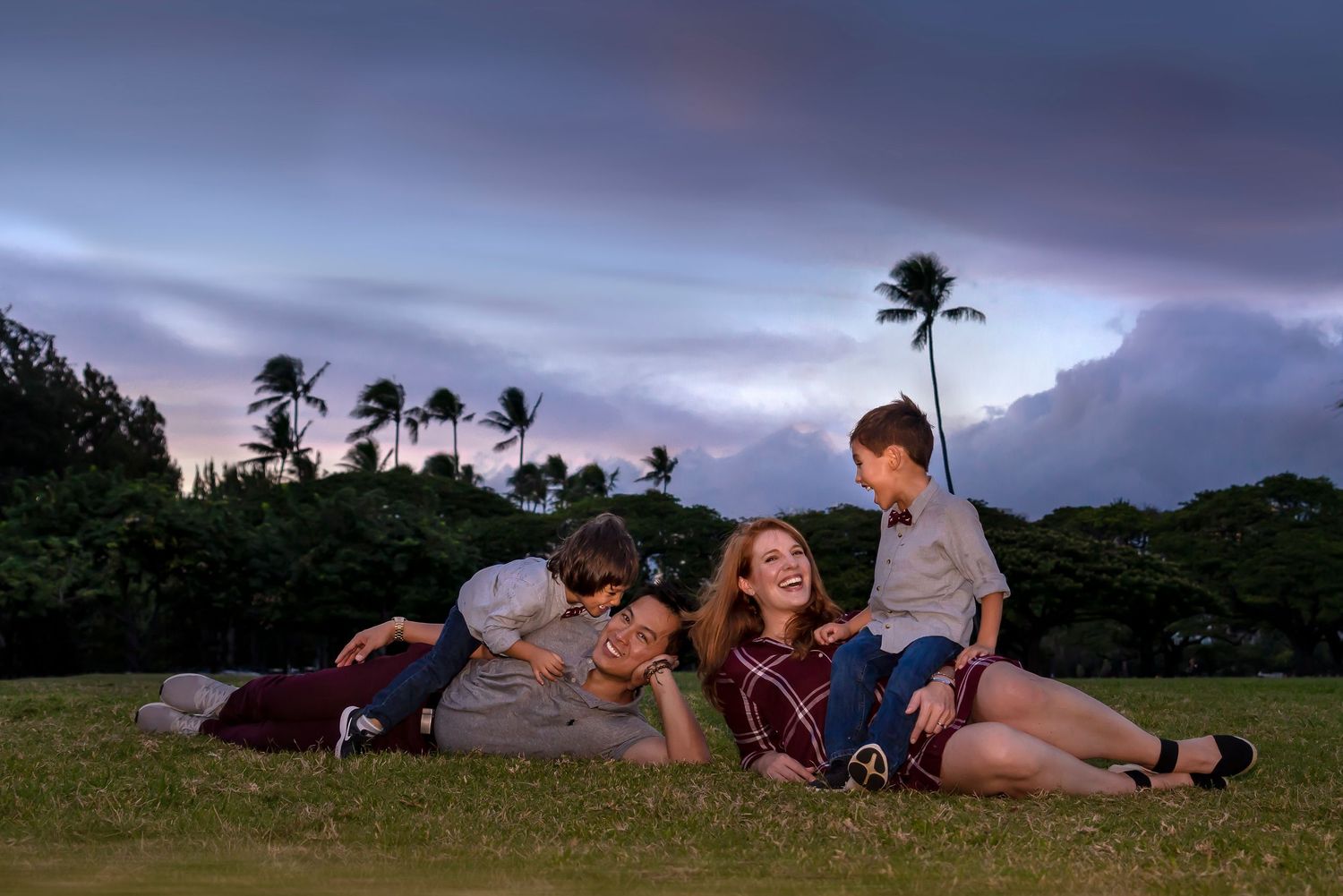 Family enjoying twilight moment on grassy hill with palm trees silhouetted against purple sky. Family Portrait Photography