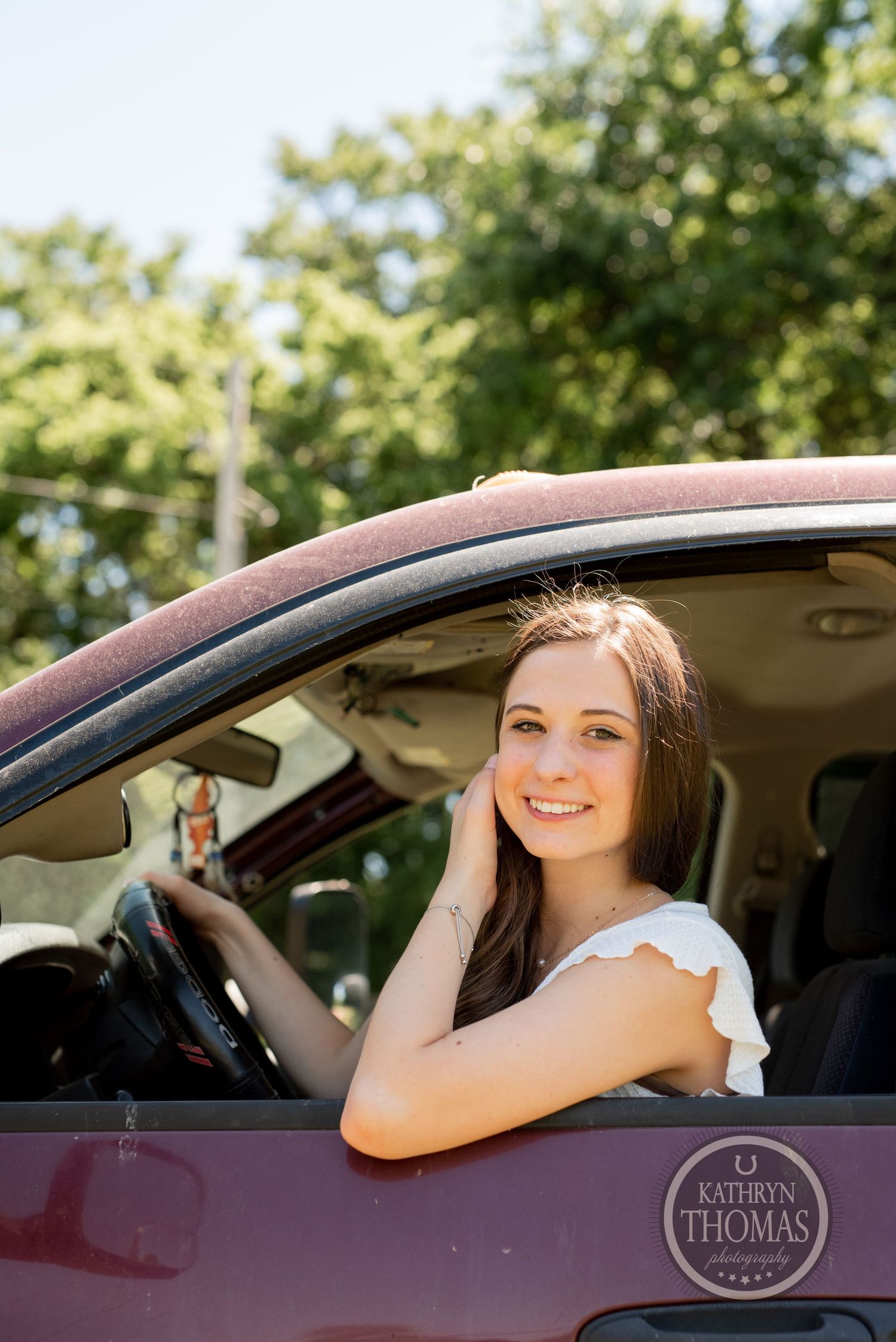 A person sitting in a car wearing a white top and smiling while resting against the window.