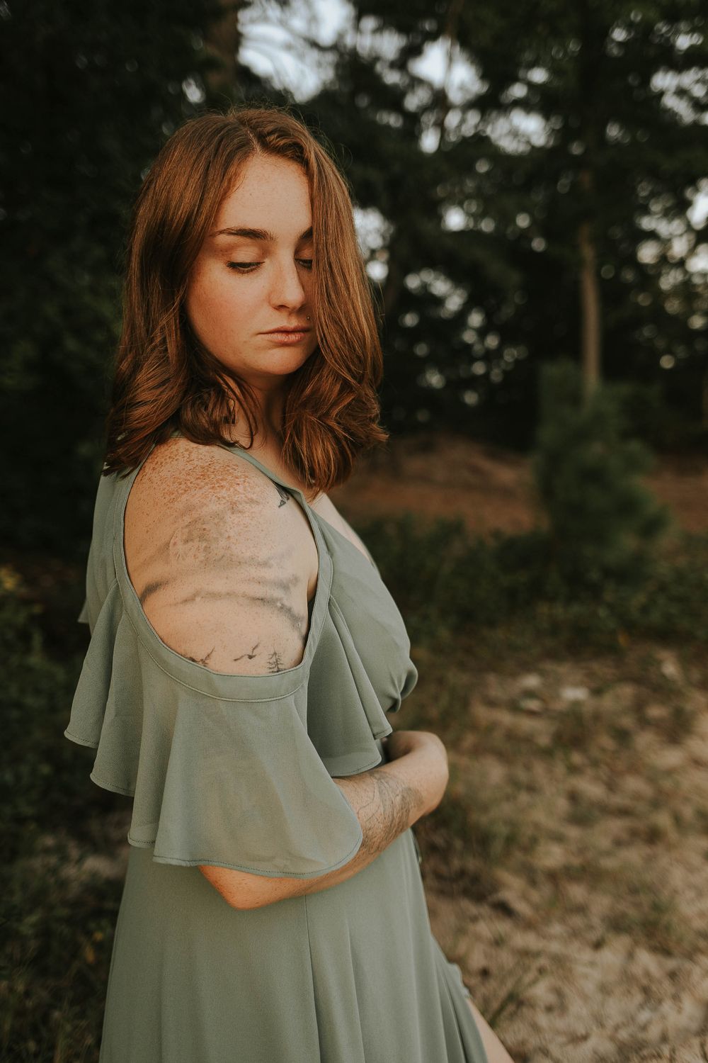 A person in a sage green dress poses thoughtfully against a natural backdrop.