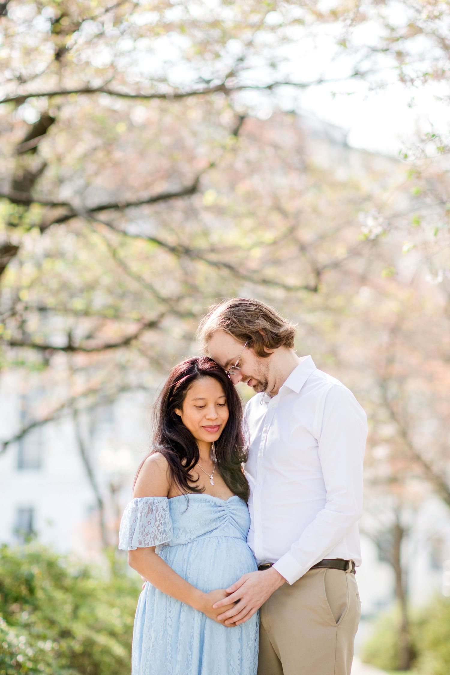 Expectant couple sharing an intimate moment under blooming spring trees.