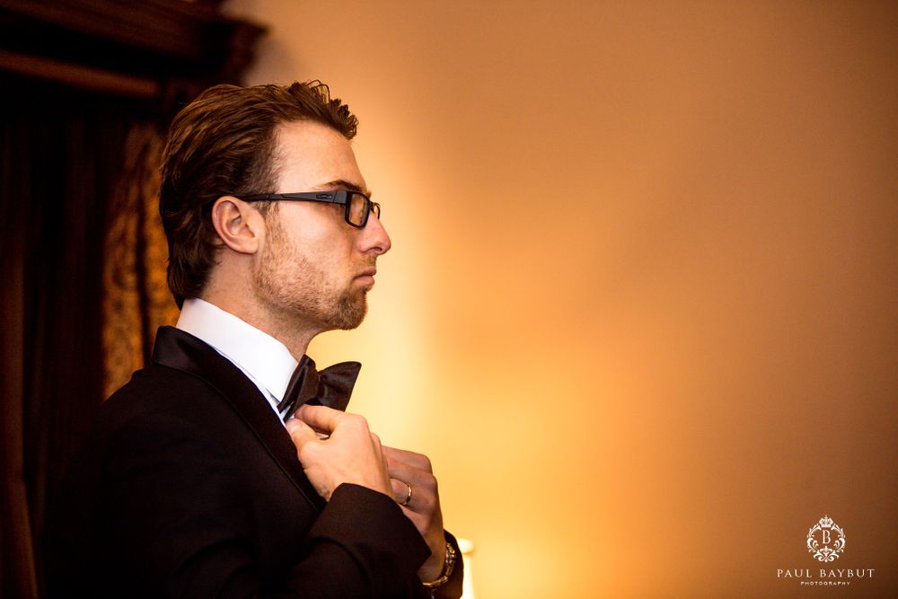 Groom in black tuxedo and glasses adjusts bowtie in warm lighting during wedding preparations.