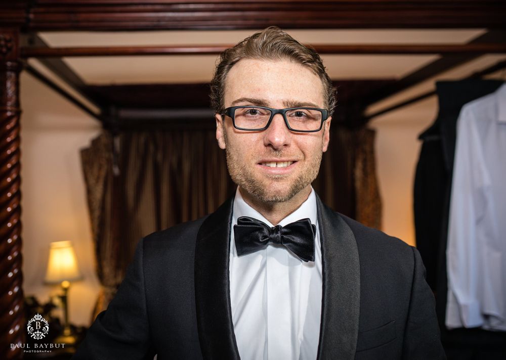 Elegant groom wearing glasses and black bow tie smiles in formal wedding attire.
