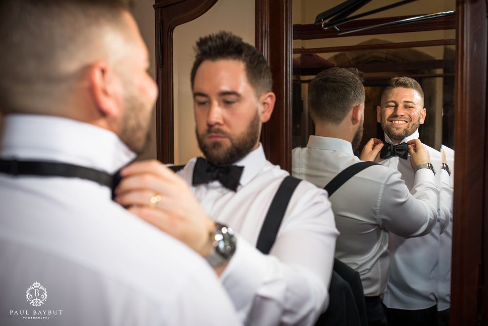 Two people adjust formal wear while looking in mirror during wedding preparations.