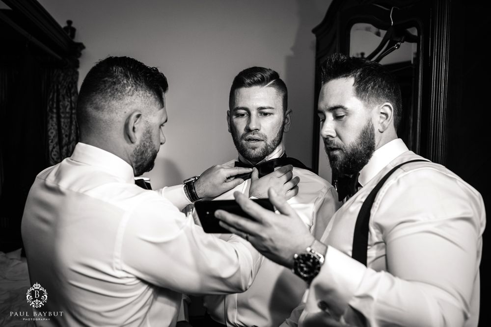 Three people in formal attire help with bow tie adjustments in black and white.