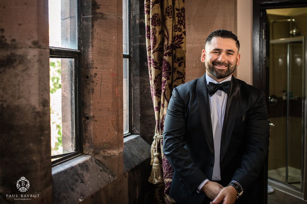 A groom in a classic black tuxedo and bowtie stands by a window during wedding preparations.