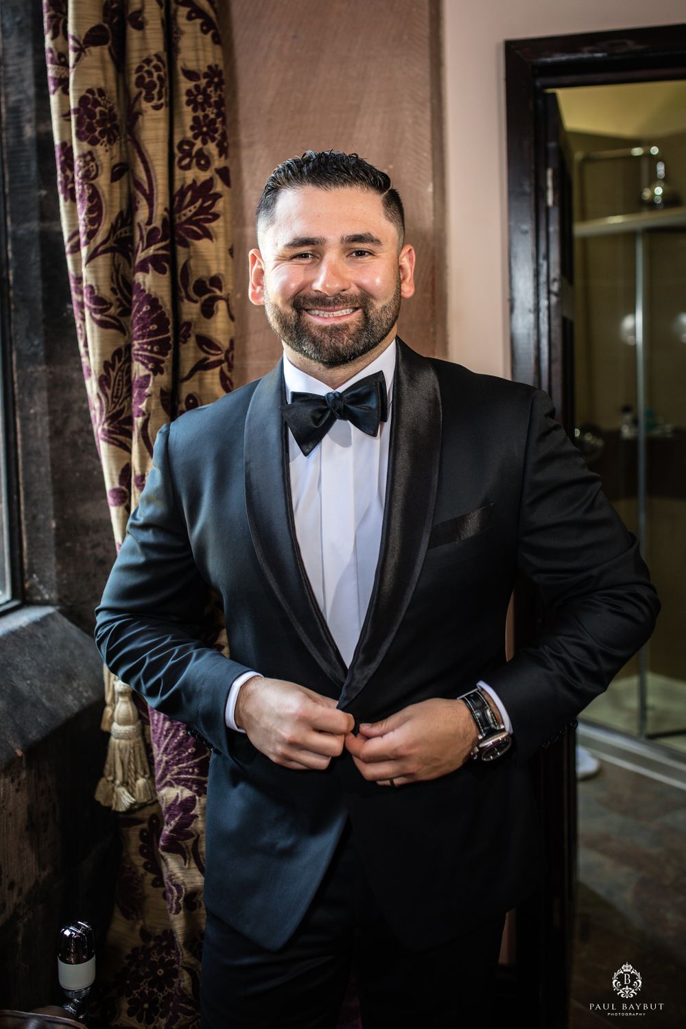 A groom adjusts his black bowtie while wearing a classic tuxedo at an elegant venue.